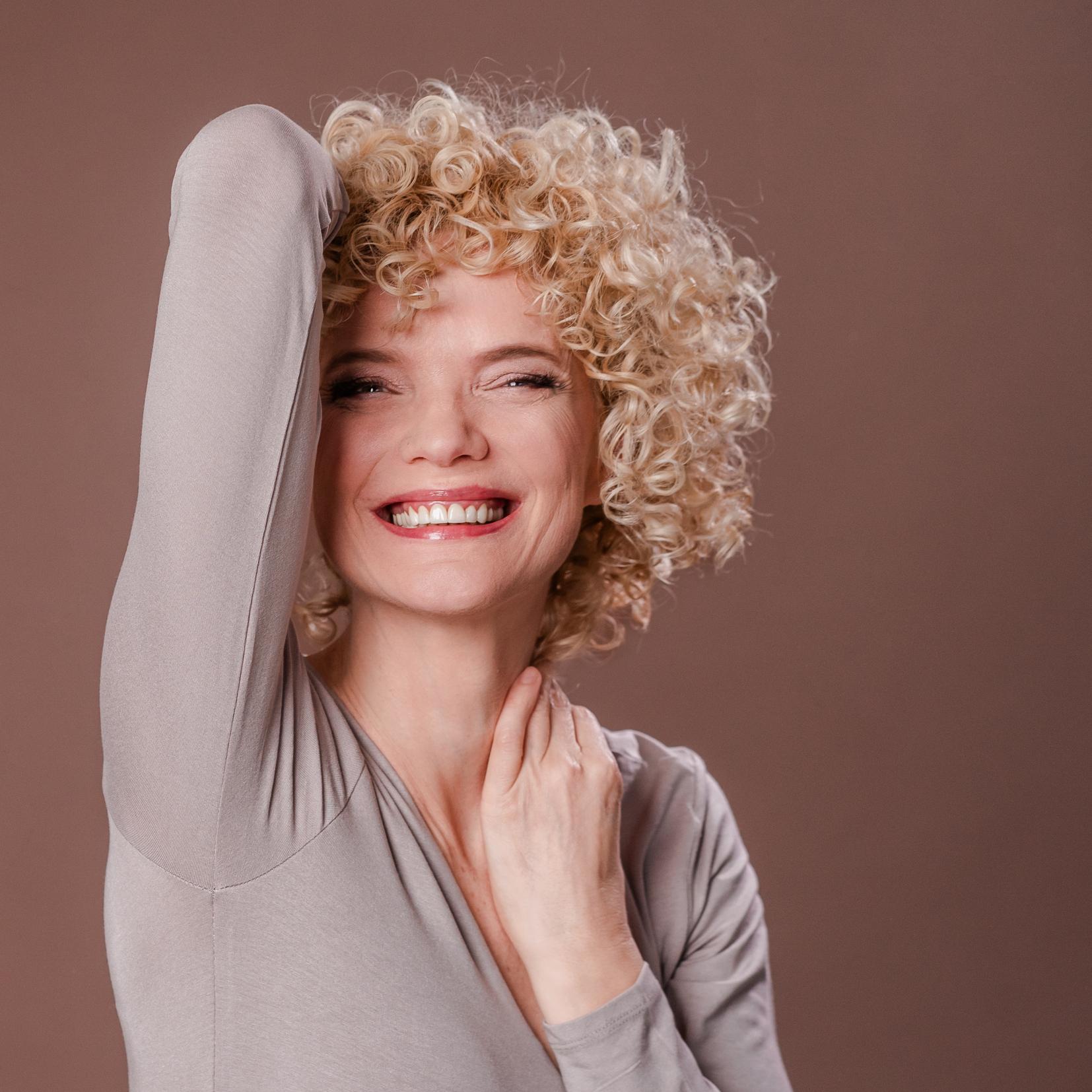 Frau mit kurzem, blondem Lockenhaarschnitt lacht und hebt entspannt den Arm